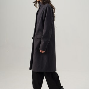 May include: A charcoal gray coat with a high collar and a single-button closure. The coat is paired with black pants and black platform boots. A tan fedora and sunglasses complete the outfit. The overall style is modern and chic.