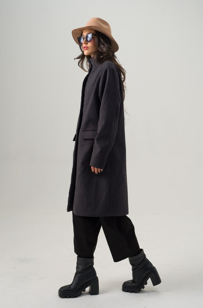 May include: A charcoal gray coat with a high collar and a single-button closure. The coat is paired with black pants and black platform boots. A tan fedora and sunglasses complete the outfit. The overall style is modern and chic.