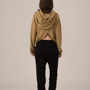 K&ouml;nnte beinhalten: Eine Person tr&auml;gt einen beigefarbenen Kapuzenpullover mit einem einzigartigen gekreuzten R&uuml;ckendesign, kombiniert mit einer schwarzen strukturierten Hose und schwarzen Schuhen. Das Outfit ist schlicht und l&auml;ssig.