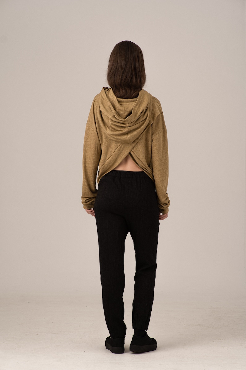 K&ouml;nnte beinhalten: Eine Person tr&auml;gt einen beigefarbenen Kapuzenpullover mit einem einzigartigen gekreuzten R&uuml;ckendesign, kombiniert mit einer schwarzen strukturierten Hose und schwarzen Schuhen. Das Outfit ist schlicht und l&auml;ssig.