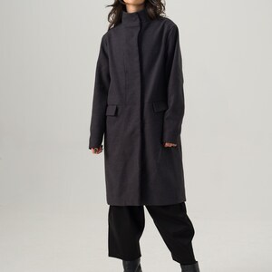 May include: A charcoal gray coat with a high collar and two front pockets. The coat is worn with black pants, black boots, a tan hat, and sunglasses. The coat is knee-length and has a simple, tailored design.