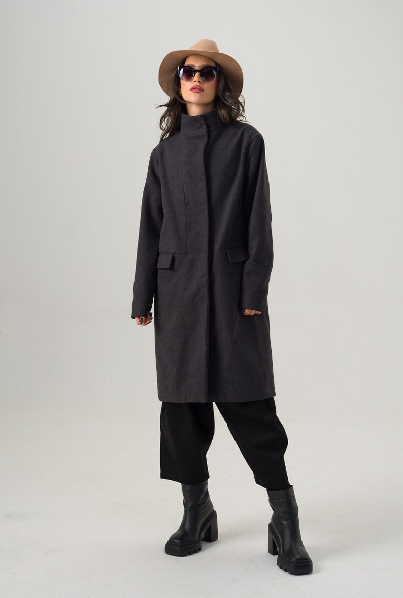 May include: A charcoal gray coat with a high collar and two front pockets. The coat is worn with black pants, black boots, a tan hat, and sunglasses. The coat is knee-length and has a simple, tailored design.