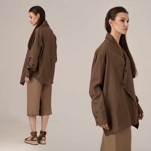 以下が含まれることがあります： 長袖とユニークな非対称デザインの、茶色のオーバーサイズのボタンダウンシャツ。シャツは、同様の色合いの膝丈のショーツと組み合わせています。茶色のサンダルでコーディネートを完成させています。