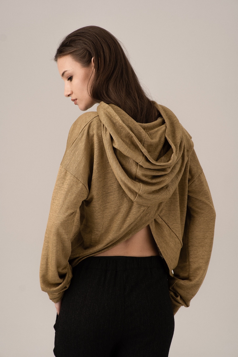 K&ouml;nnte beinhalten: Ein senffarbener Kapuzenpullover mit einem einzigartigen R&uuml;ckendesign, kombiniert mit einer schwarzen Hose. Der Kapuzenpullover hat eine drapierte Kapuze und lange &Auml;rmel. Das Model ist von hinten zu sehen, wobei der R&uuml;cken des Hoodies und die Taille hervorgehoben werden.