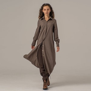 May include: A long brown shirt dress with a button-up front and long sleeves. The dress has a slightly asymmetrical hemline and is worn with brown pants and brown boots.