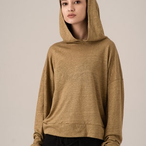 K&ouml;nnte beinhalten: Ein beigefarbener Kapuzenpullover mit langen &Auml;rmeln. Der Pullover hat eine lockere Passform und einen Kordelzug am unteren Saum. Das Model tr&auml;gt eine schwarze Hose. Der Stoff scheint ein leichter Strick zu sein.