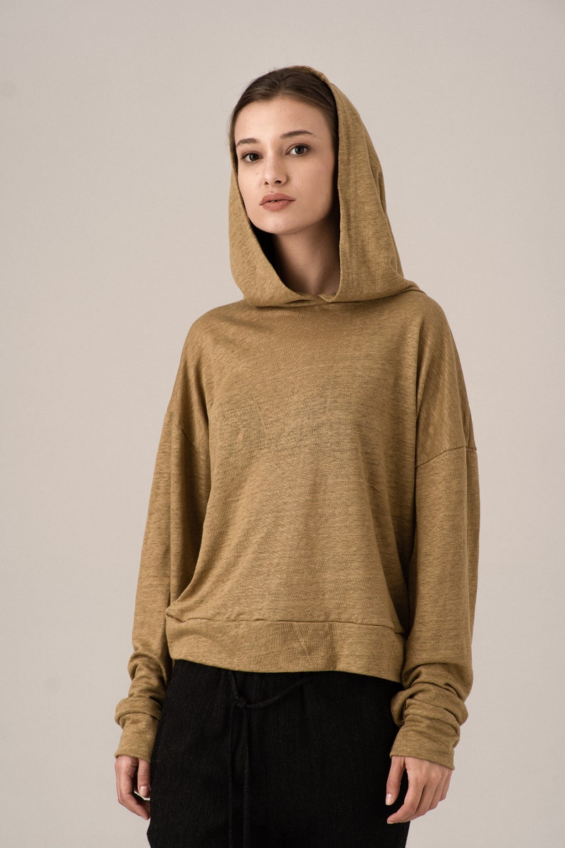 K&ouml;nnte beinhalten: Ein beigefarbener Kapuzenpullover mit langen &Auml;rmeln. Der Pullover hat eine lockere Passform und einen Kordelzug am unteren Saum. Das Model tr&auml;gt eine schwarze Hose. Der Stoff scheint ein leichter Strick zu sein.