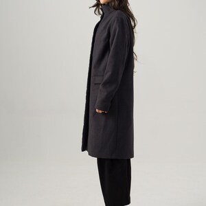 May include: A dark gray, knee-length coat with a high collar and two front pockets. The coat is worn with black pants, black boots, a tan hat, and sunglasses. The outfit is a stylish ensemble.