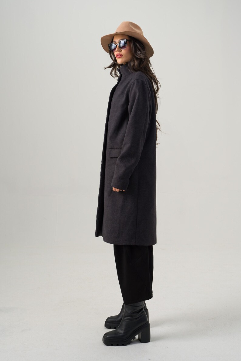 May include: A dark gray, knee-length coat with a high collar and two front pockets. The coat is worn with black pants, black boots, a tan hat, and sunglasses. The outfit is a stylish ensemble.