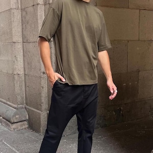 Puede incluir: Una persona con una camiseta verde oscuro de gran tamaño, pantalones negros y zapatos negros.