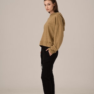 K&ouml;nnte beinhalten: Eine Frau tr&auml;gt einen beigefarbenen Kapuzenpullover und eine schwarze Hose. Der Kapuzenpullover hat eine Kapuze mit Kordelzug und lange &Auml;rmel. Die schwarze Hose ist locker geschnitten und hat einen Kordelzug in der Taille. Schwarze Turnschuhe vervollst&auml;ndigen das Outfit.