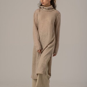 May include: A long, beige knitted sweater dress with a turtleneck and side slits. The dress is worn with beige pants and brown ankle boots.