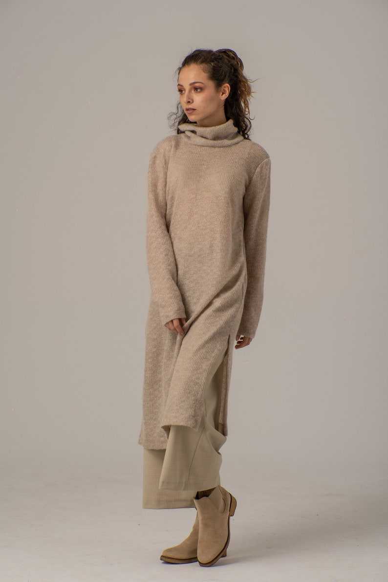 May include: A long, beige knitted sweater dress with a turtleneck and side slits. The dress is worn with beige pants and brown ankle boots.