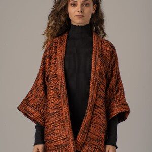 Op de afbeelding: Een vrouw draagt een langarmige, oranje en zwarte gebreide cardigan met een zwarte turtleneck eronder.