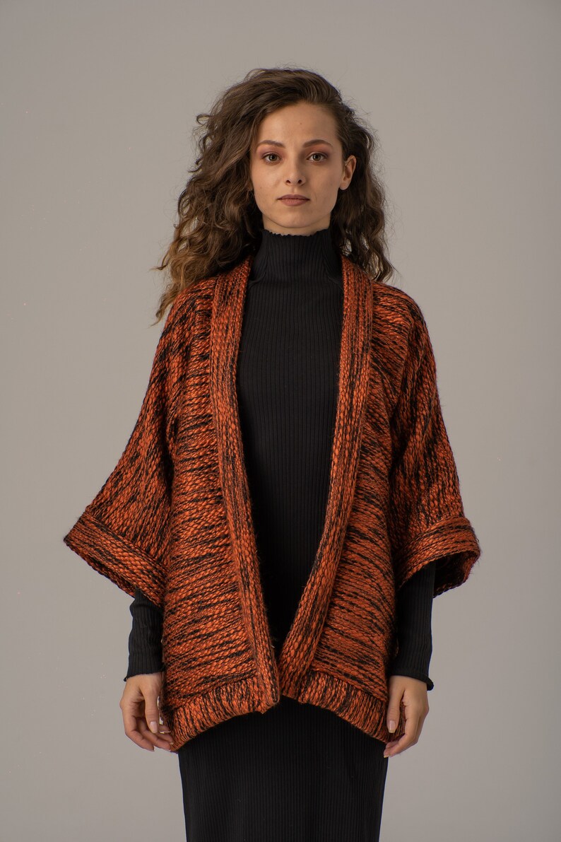 Op de afbeelding: Een vrouw draagt een langarmige, oranje en zwarte gebreide cardigan met een zwarte turtleneck eronder.