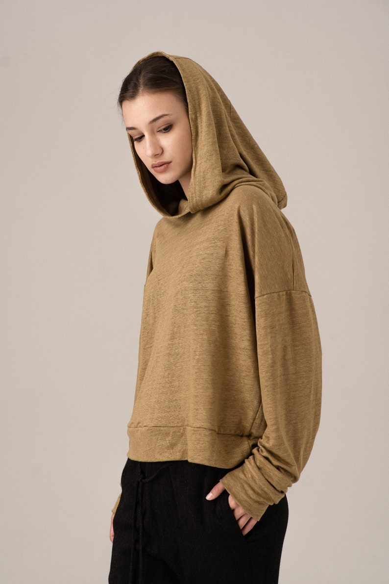 K&ouml;nnte beinhalten: Ein olivgr&uuml;nes, kurz geschnittenes Kapuzensweatshirt. Das lang&auml;rmlige Oberteil hat eine lockere Passform und einen Kordelzug am Saum. Das Model tr&auml;gt eine schwarze Hose.