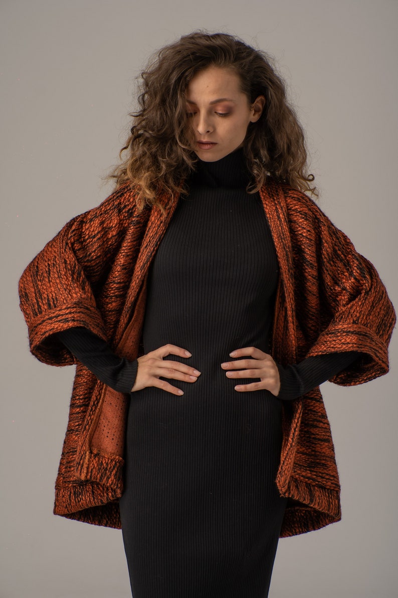 Op de afbeelding: Een vrouw draagt een zwarte turtleneck trui en een lange, oranje en zwarte gepatroneerde cardigan. De cardigan heeft een losse pasvorm en lange mouwen.