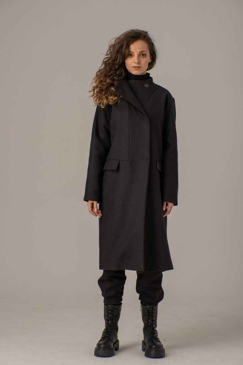 May include: A black long-sleeved coat with a high collar and a single button closure. The coat is worn by a person with long curly hair. The person is wearing black pants and black boots.