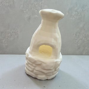 May include: A white, abstract ceramic sculpture resembling a stylized cave or shelter. The sculpture has a textured, layered base and a rounded opening in the center, with a soft yellow glow emanating from within. The top has a unique, irregular shape.