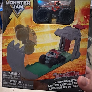 May include: A Monster Jam Thunder Roarus launcher playset. The box features a monster truck toy and a launcher set with a dinosaur-themed design. The toy truck is red and grey with large black tyres. The packaging includes warning labels.