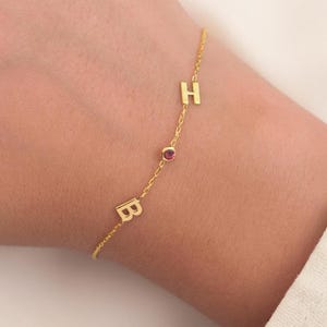 May include: A fine gold bracelet featuring the letters "B" and "H" and a small red gemstone. The bracelet is worn on a wrist, set against a soft, neutral background. This is a personalized piece of jewellery.