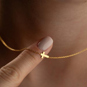May include: A delicate gold-colored necklace featuring a small cross pendant. The cross is positioned to the side, suspended from a fine chain. The necklace is displayed against a blurred background.
