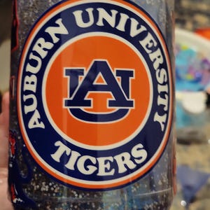 May include: A clear glass jar filled with blue glitter. The jar features a circular emblem with the words "Auburn University Tigers" in navy blue and orange. The emblem includes a stylized letter "A".