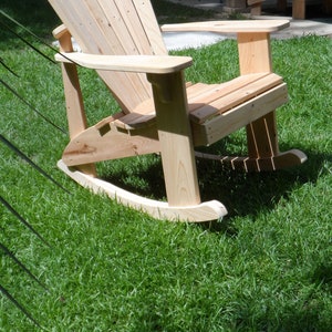 May include: A wooden rocking chair with a slatted back and arms. The chair is made of natural wood and has a rustic look. It is sitting on a green grassy lawn.