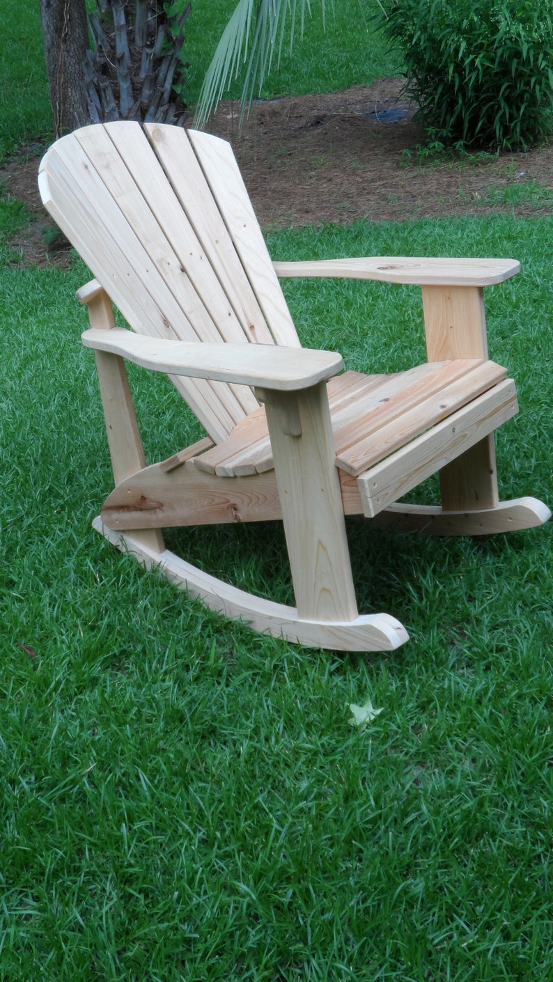 May include: A wooden rocking chair with a high back and wide arms. The chair is made of natural wood and has a rustic look. It is sitting on a green grassy lawn.