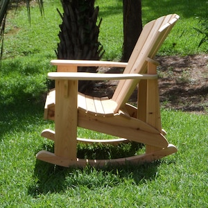 May include: A wooden rocking chair with a natural finish. The chair is made of slats and has a high back. It is sitting on a grassy lawn.