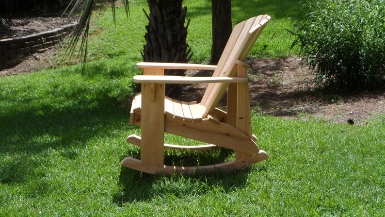 May include: A wooden rocking chair with a natural finish. The chair is made of slats and has a high back. It is sitting on a grassy lawn.