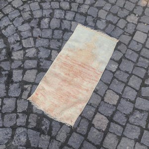 May include: A light pink and white vintage rug with a faded design. The rug is fringed on all sides and has a worn, distressed look.