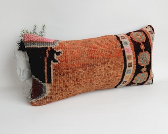 May include: A long, rectangular pillow with a brown, orange, and black patterned fabric. The pillow has a geometric design with a border of small, colorful squares and circles.