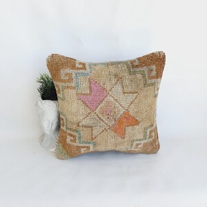 May include: Square decorative pillow with a geometric pattern. The pillow features a beige background with a central star design in pink and orange, surrounded by brown and blue accents. The pillow is placed on a white surface.
