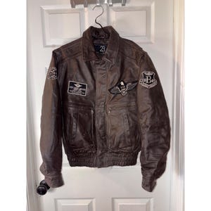 May include: A dark brown leather jacket featuring a fold-down collar, ribbed cuffs, and a ribbed hem. The front is decorated with multiple embroidered patches, including a black and white wing emblem with the text F19 ARMY and a circular B logo.