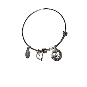 May include: Silver-toned bangle bracelet with three charms. Charms include a small oval tag, a simple heart, and a heart-shaped pendant with the word "FOREVER" and a small gem. The bracelet is on a white background.