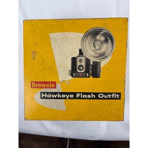 May include: A square yellow cardboard box for a Brownie Hawkeye Flash Outfit. The box features a graphic illustration of a black camera with a large circular silver flash attachment. The text reads Brownie and Hawkeye Flash Outfit in white.