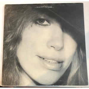 Puede incluir: Portada de álbum en blanco y negro con un primer plano del rostro de una persona, parcialmente cubierto por un sombrero. La persona sonríe, con pelo largo y una mirada directa. El título del álbum y el nombre del artista son visibles.