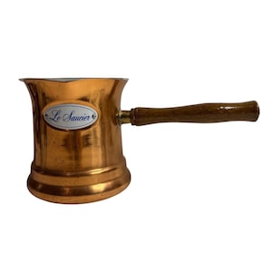 May include: A copper sauce pot with a flared rim and a long, straight wooden handle. The pot features an oval white enamel plaque on the front with the blue text Le Saucier. The metallic surface has a warm, polished finish with visible texture.