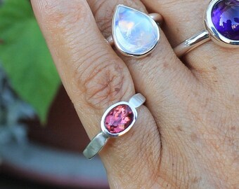 Könnte beinhalten: Zwei Silberringe mit Edelsteinen. Der Ring links hat einen tropfenförmigen Mondstein. Der Ring rechts hat einen rosafarbenen ovalen Edelstein.