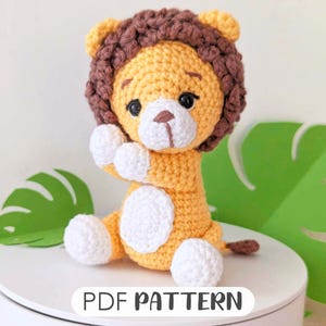 Peut inclure: Un jouet en crochet amigurumi en forme de lion avec une crinière brune, un corps jaune et un visage blanc. Le jouet est assis sur une surface blanche avec une feuille verte derrière lui. Le texte "PDF PATTERN" est imprimé sous le jouet.