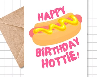 May include: A white greeting card with a cartoon hot dog design, mustard, and the text "HAPPY BIRTHDAY HOTTIE!" in pink. A brown envelope is partially visible in the background.