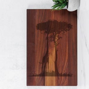 Könnte beinhalten: Rechteckiges Holzschneidebrett mit einer Giraffensilhouette unter einem Baum. Die Giraffe und der Baum sind in das Holz geätzt und bilden einen dunklen Kontrast zum helleren Holzmaserung. Das Brett hat eine natürliche Holzoberfläche.