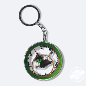 May include: A circular keyring with a green border and a clear centre, depicting a cartoon witch in a pointed hat. The keyring has a black ring and a silver chain. Small black heart-shaped accents surround the witch.