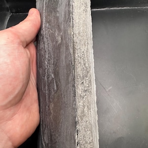 May include: A rectangular, dark gray stone slab with a rough, textured edge. The stone is held by a hand, showcasing its size and the natural variations in its surface. The slab appears to be approximately 10 inches long.