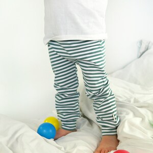 May include: A child wearing green and white striped pants stands on a white bed with colorful balls scattered around.