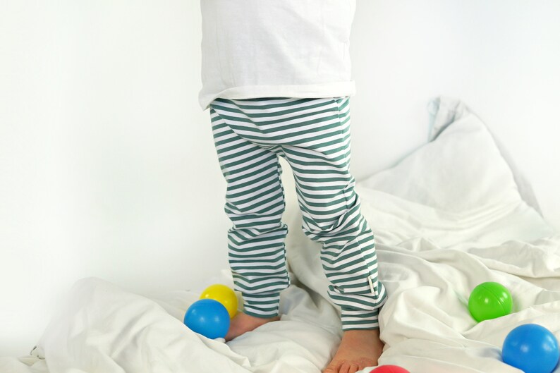 May include: A child wearing green and white striped pants stands on a white bed with colorful balls scattered around.