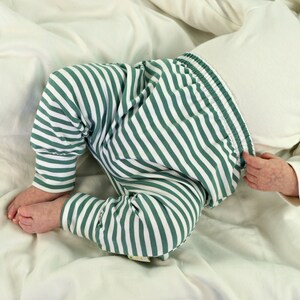 May include: A baby wearing green and white striped pants with a white long-sleeved shirt. The baby is lying on a white sheet.