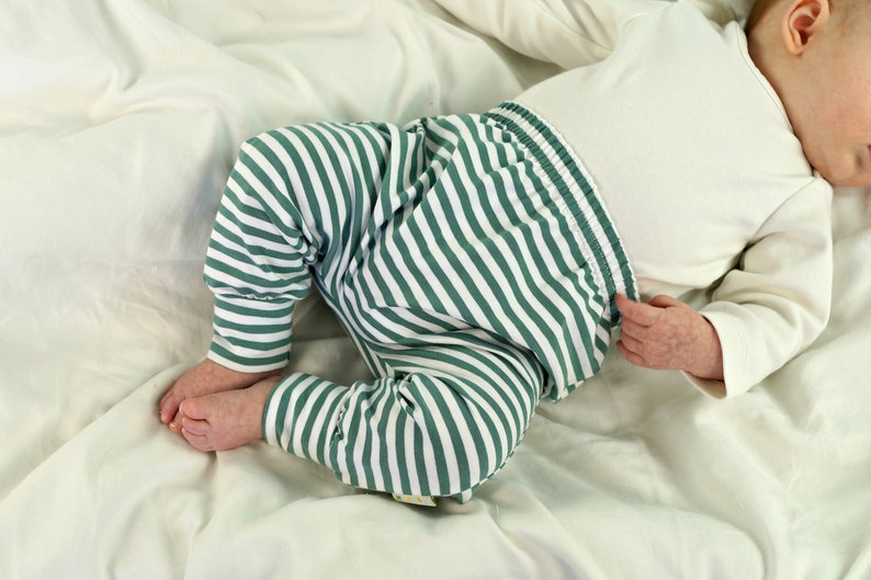 May include: A baby wearing green and white striped pants with a white long-sleeved shirt. The baby is lying on a white sheet.