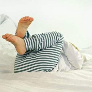 May include: A child wearing green and white striped pajamas lies on a white bed with their feet in the air.